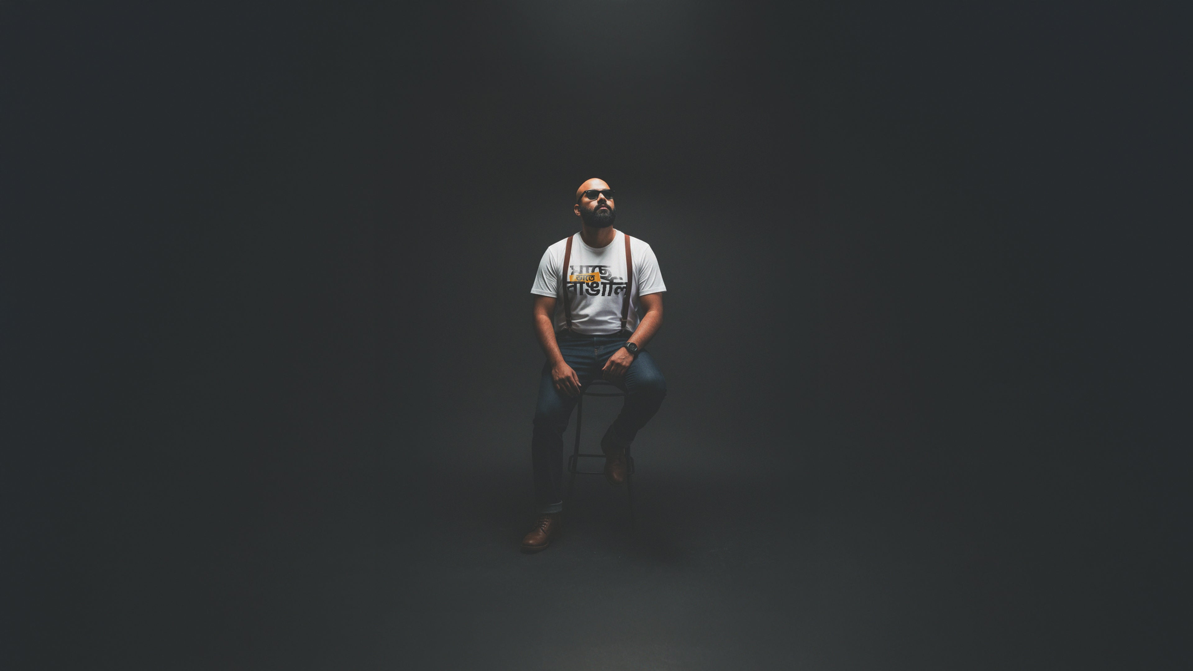Man sitting on a chair in a dark room wearing a white t-shirt with text. Bangla streetwear Bengali street wear t-shirt